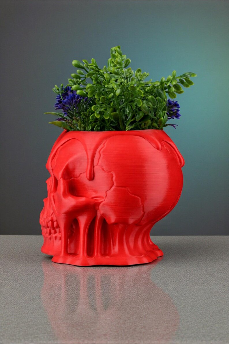 May include: A red skull-shaped planter with a textured surface, holding green and purple plants. The planter has a unique design with a dripping effect around the top. A decorative item for home decor.