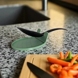 May include: A green silicone spoon rest with a black spoon resting on it. The spoon rest is on a speckled countertop next to a wooden cutting board with a knife and cut vegetables.
