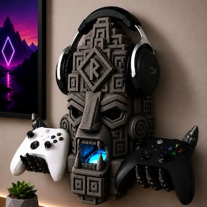 Könnte beinhalten: Ein wandmontierter Gaming-Organizer in grauer Steinoptik, geformt wie eine geschnitzte Maske mit leuchtend blauem Mund. Er verfügt über zwei klauenartige Hände, die einen weißen und einen schwarzen Controller halten, sowie einen schwarzen Kopfhörer.