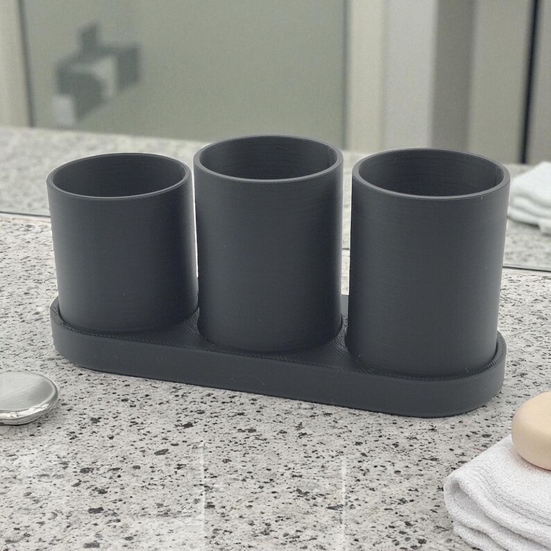 May include: A dark grey bathroom organiser featuring three cylindrical holders. The organiser sits on a rectangular base, designed for toothbrushes, toothpaste, or other bathroom essentials. The minimalist design is suitable for any bathroom decor.