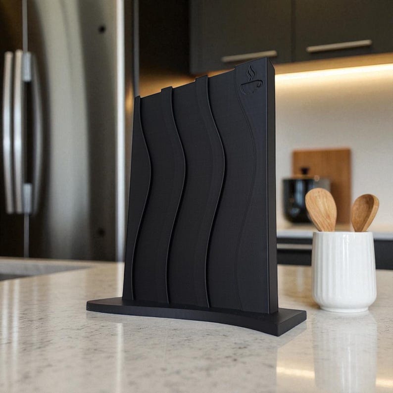 May include: A black knife block with a wavy design. The block has a rectangular base and a curved top. A small coffee cup icon is visible on the top right corner. The block is on a white countertop. A white ceramic container with wooden spoons is to the right.
