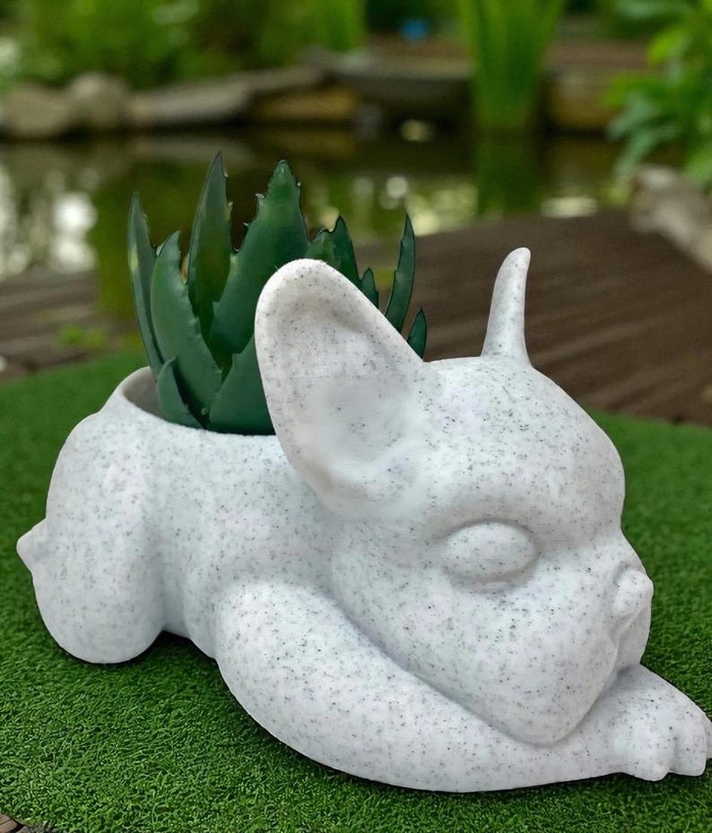 May include: A white, speckled ceramic planter in the shape of a French bulldog. The dog is lying down with its head resting on its paws. The planter holds a green succulent plant. The background is blurred, showing a green lawn and a pond.