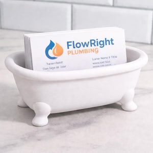 May include: A white, miniature bathtub-shaped business card holder. The holder is filled with white business cards. The cards display the "FlowRight Plumbing" logo and contact information. The holder sits on a marble surface.