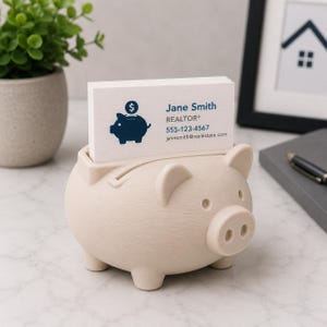 May include: A beige piggy bank-shaped business card holder. The holder has a slot on top and holds a stack of white business cards. The business cards have a blue piggy bank logo and the name "Jane Smith, REALTOR&reg;" with contact information.