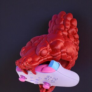 May include: A red, textured sculpture of a snake's head biting a white, pink, and blue video game controller. The controller has pink buttons and a blue accent. The snake's tongue is red.