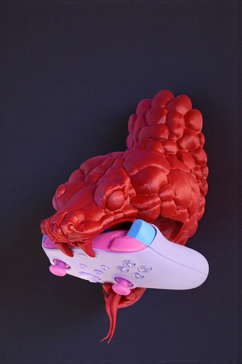 May include: A red, textured sculpture of a snake's head biting a white, pink, and blue video game controller. The controller has pink buttons and a blue accent. The snake's tongue is red.