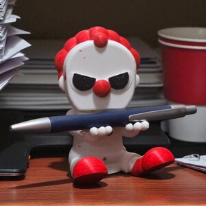 May include: A white and red clown figurine holding a blue pen with a silver tip. The clown has a red nose, red hair, and black eyes. The figurine is sitting on a wooden surface.