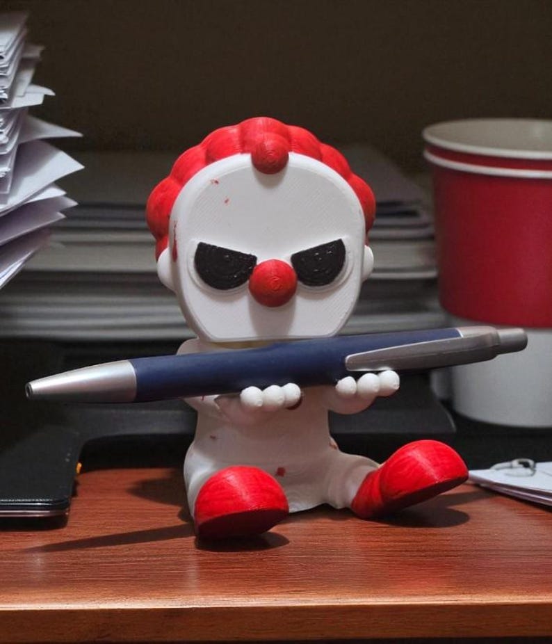 May include: A white and red clown figurine holding a blue pen with a silver tip. The clown has a red nose, red hair, and black eyes. The figurine is sitting on a wooden surface.
