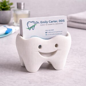 Op de afbeelding: Een witte, tandvormige visitekaarthouder met een lachend gezicht. De houder bevat visitekaartjes voor "Dr. Emily Carter, DDS, Family & Cosmetic Dentistry". De kaarten hebben een blauw en groen logo. De houder staat op een lichtgekleurd oppervlak.