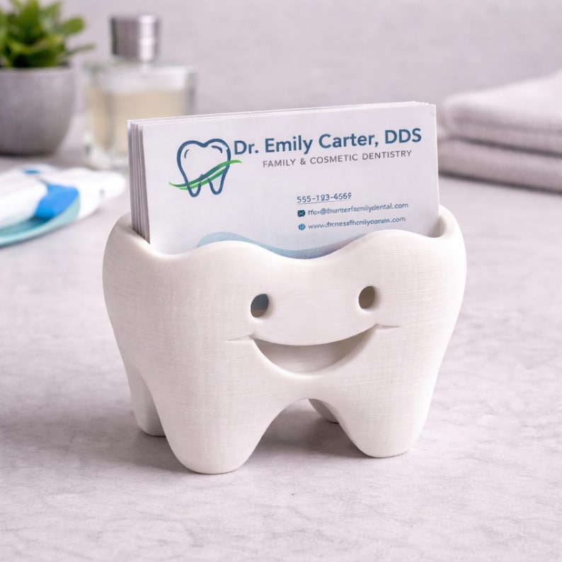 Op de afbeelding: Een witte, tandvormige visitekaarthouder met een lachend gezicht. De houder bevat visitekaartjes voor "Dr. Emily Carter, DDS, Family & Cosmetic Dentistry". De kaarten hebben een blauw en groen logo. De houder staat op een lichtgekleurd oppervlak.