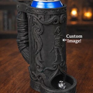 May include: Black, intricately carved can holder with a built-in dice tower. The holder features a detailed relief of a figure and scrollwork. A blue can sits in the top. The text "Custom Image!" is visible.