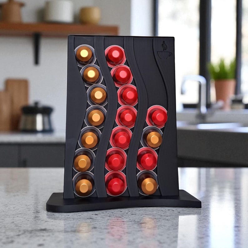 May include: A black coffee pod holder with a curved design, holding various colored coffee pods. The holder has a base and a coffee cup icon. The pods are in shades of gold and red.