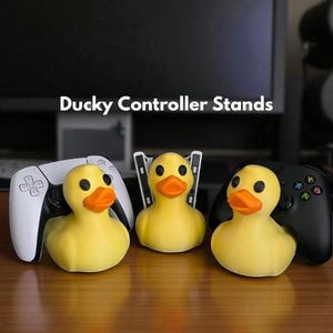 Peut inclure: Trois supports de manette en forme de canard en caoutchouc jaune avec des becs orange et des yeux noirs. Chaque canard tient une manette de jeu différente : une PlayStation 5 blanche, une Xbox noire et une manette blanche. Le texte "Ducky Controller Stands" est en haut.
