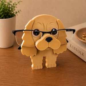 K&ouml;nnte beinhalten: Ein brillenf&ouml;rmiger Halter in Form eines beigefarbenen Hundes mit einer schwarzen Brille. Der Halter hat gro&szlig;e, ausdrucksstarke Augen und eine schwarze Nase. Die Brille liegt auf der Nase des Hundes. Eine kleine Topfpflanze und ein Buch sind im Hintergrund.