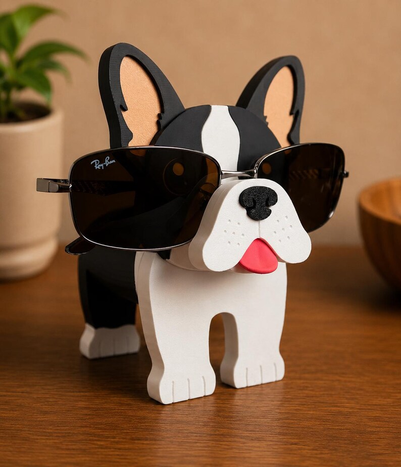 May include: A novelty sunglasses holder shaped like a French bulldog. The black and white dog has a red tongue and is wearing Ray-Ban sunglasses. The holder is made of wood and sits on a wooden surface.