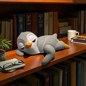 Pu&ograve; includere: Una statuetta di pinguino grigio con becco arancione e piedi gialli &egrave; sdraiata su una mensola di legno. Il pinguino ha gli occhi chiusi, suggerendo che stia dormendo. Libri e una piccola pianta sullo sfondo.