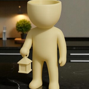 K&ouml;nnte beinhalten: Hellgelbe Figur in Form einer Person mit gro&szlig;em, offenem Kopf, die eine kleine Laterne h&auml;lt. Die Figur steht auf einer schwarz-wei&szlig;en Marmoroberfl&auml;che. Der Hintergrund umfasst eine kleine Pflanze und dunkle Schr&auml;nke.