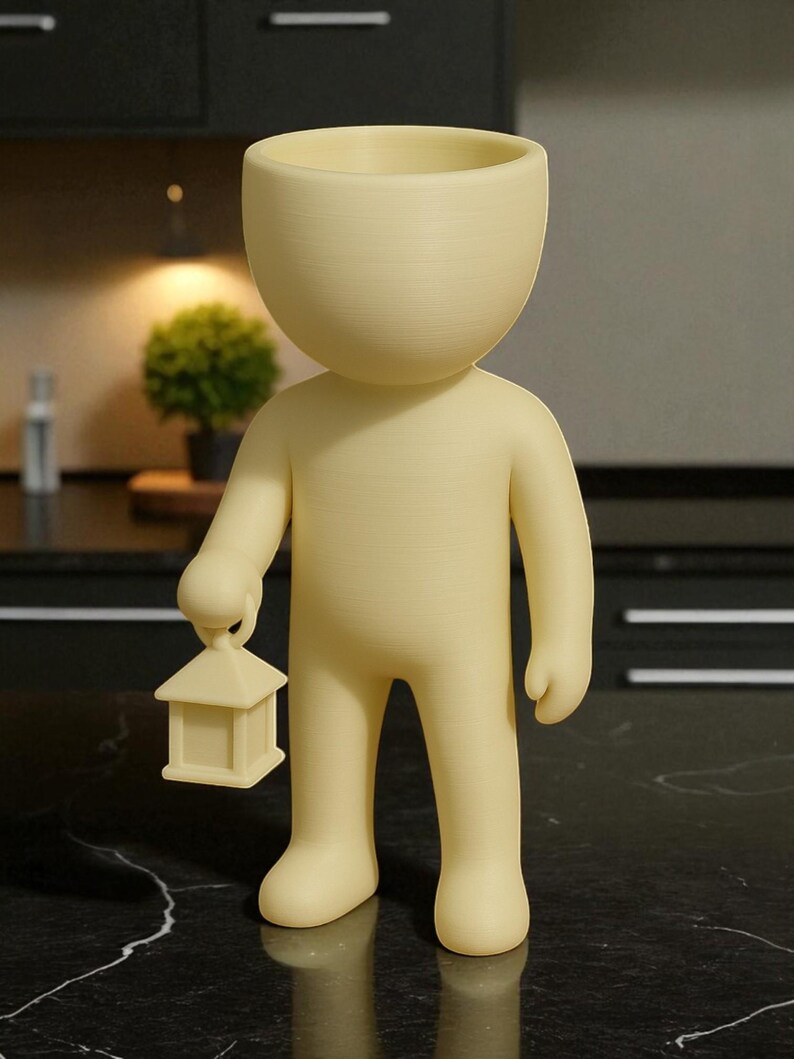 K&ouml;nnte beinhalten: Hellgelbe Figur in Form einer Person mit gro&szlig;em, offenem Kopf, die eine kleine Laterne h&auml;lt. Die Figur steht auf einer schwarz-wei&szlig;en Marmoroberfl&auml;che. Der Hintergrund umfasst eine kleine Pflanze und dunkle Schr&auml;nke.