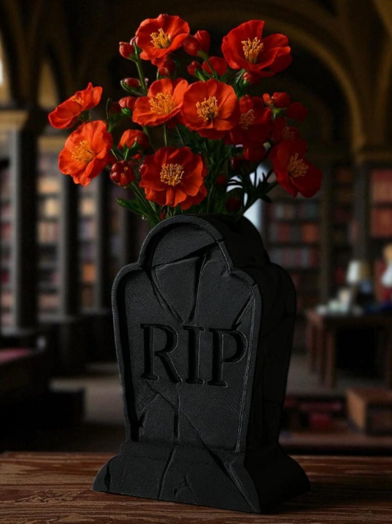 May include: A black tombstone-shaped vase with the letters "RIP" carved into the front, holding a bouquet of vibrant orange flowers with yellow centers. The vase is on a wooden surface.