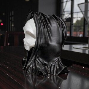 May include: A 3D-printed sculpture of a skull wearing a black hooded cloak. The skull is white, and the cloak is a glossy black. The sculpture is on a dark wooden surface. The overall theme is gothic or Halloween.