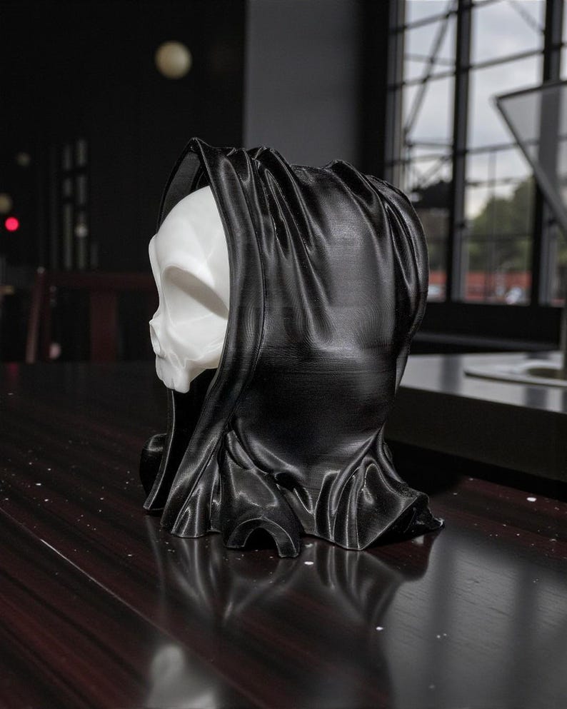 May include: A 3D-printed sculpture of a skull wearing a black hooded cloak. The skull is white, and the cloak is a glossy black. The sculpture is on a dark wooden surface. The overall theme is gothic or Halloween.