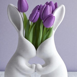 May include: A white ceramic bunny vase holding a bouquet of purple tulips. The vase is shaped like two bunnies embracing, with their ears forming the vase's opening. The tulips have green stems and leaves, set against a soft purple background.