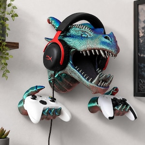 Peut inclure: Une sculpture murale représentant une tête de dragon aux écailles bleu et vert iridescentes, avec une gueule ouverte et des dents pointues. Le dragon porte un casque audio noir et rouge et ses deux mains griffues tiennent chacune une manette blanche.