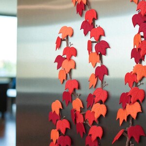 May include: Decorative fall leaf garlands in shades of red and orange. The leaves are attached to brown branches, creating a seasonal autumn theme. The garlands are arranged vertically, adding a touch of nature-inspired decor.