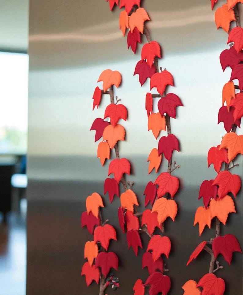 May include: Decorative fall leaf garlands in shades of red and orange. The leaves are attached to brown branches, creating a seasonal autumn theme. The garlands are arranged vertically, adding a touch of nature-inspired decor.