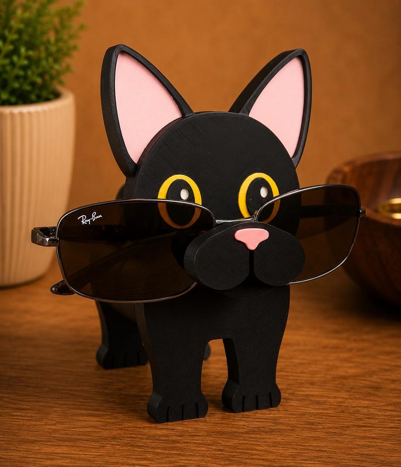 May include: A black wooden dog-shaped eyeglass holder with pink ears and a pink nose. The dog is wearing a pair of sunglasses with dark lenses. The holder is on a wooden surface.