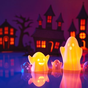May include: Five glowing white ghost figurines with orange lights inside, sitting in front of a dark silhouette of a haunted house with lit windows.
