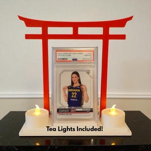 May include: A red torii gate-shaped display stand holding a Caitlin Clark basketball card. Two battery-operated tea lights sit on a white base. The card is in a protective case and features the text "2024 Panini Instant WNBA Caitlin Clark".