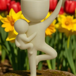 May include: A cream-colored figurine depicting a parent figure holding a child, with one arm raised and standing on one leg. The parent figure has a cup-shaped head. The background features red tulips and yellow daffodils.
