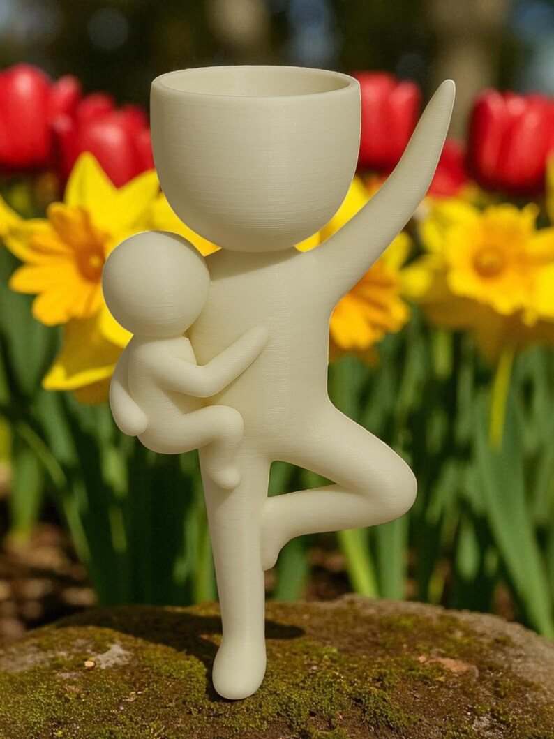 May include: A cream-colored figurine depicting a parent figure holding a child, with one arm raised and standing on one leg. The parent figure has a cup-shaped head. The background features red tulips and yellow daffodils.
