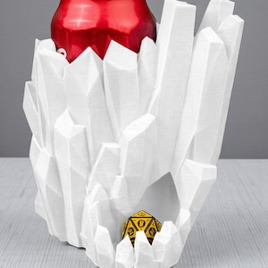 May include: A white, geometric, crystal-shaped can holder with a red soda can inside. A yellow and black polyhedral die rests in a lower cavity of the holder. The holder is made of a rigid material.