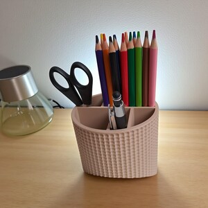 May include: A beige desk organizer with a textured basket weave design, holding colored pencils, a black pen, and scissors. The pencils are various colors, including red, green, and pink. A silver and black pen is also visible.