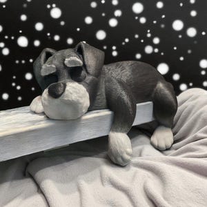 May include: A gray and white dog figurine resting on a white wooden ledge. The dog has a white muzzle, paws, and eyebrows. The background is black with white circular shapes. The dog is lying on a gray blanket.