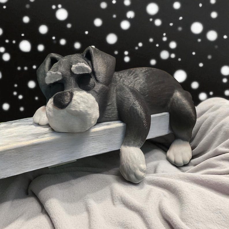 May include: A gray and white dog figurine resting on a white wooden ledge. The dog has a white muzzle, paws, and eyebrows. The background is black with white circular shapes. The dog is lying on a gray blanket.