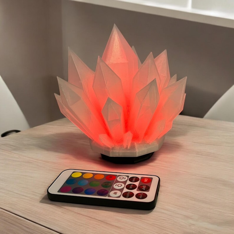 May include: A crystal-shaped lamp with a red glow, sitting on a light-colored surface. The lamp has a white base and a remote control with color options. The lamp's design resembles a cluster of crystals.