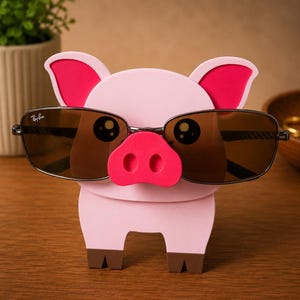 May include: A pink pig-shaped eyeglass holder with a red snout and ears. The holder is designed to hold a pair of sunglasses with brown lenses and black frames. The pig has cartoon-like eyes and brown hooves.