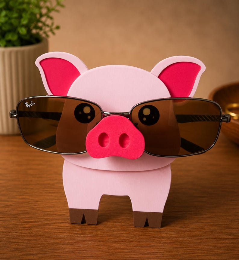 May include: A pink pig-shaped eyeglass holder with a red snout and ears. The holder is designed to hold a pair of sunglasses with brown lenses and black frames. The pig has cartoon-like eyes and brown hooves.