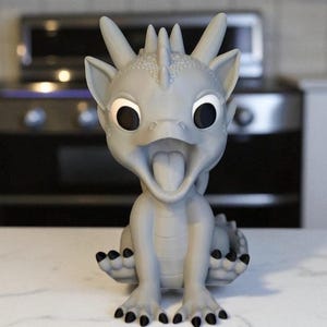 May include: A gray dragon figurine with large, expressive eyes and an open mouth, revealing a tongue. The dragon has black claws and a textured body, sitting upright. The background is blurred, suggesting a kitchen setting.