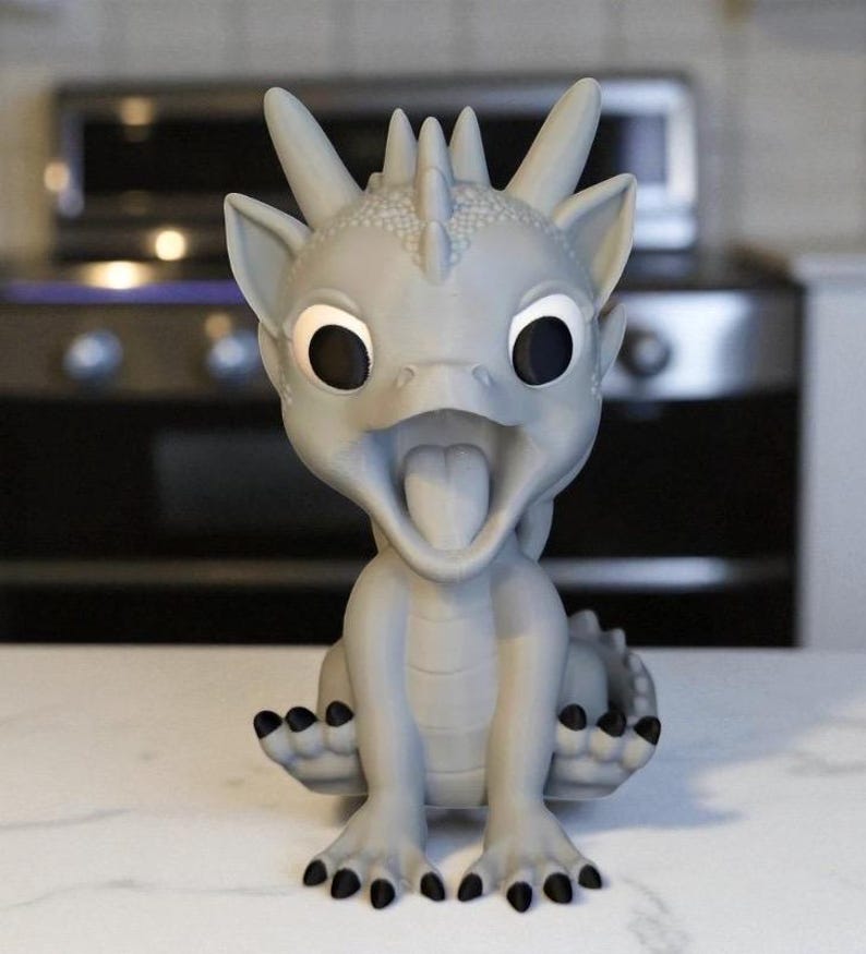 May include: A gray dragon figurine with large, expressive eyes and an open mouth, revealing a tongue. The dragon has black claws and a textured body, sitting upright. The background is blurred, suggesting a kitchen setting.