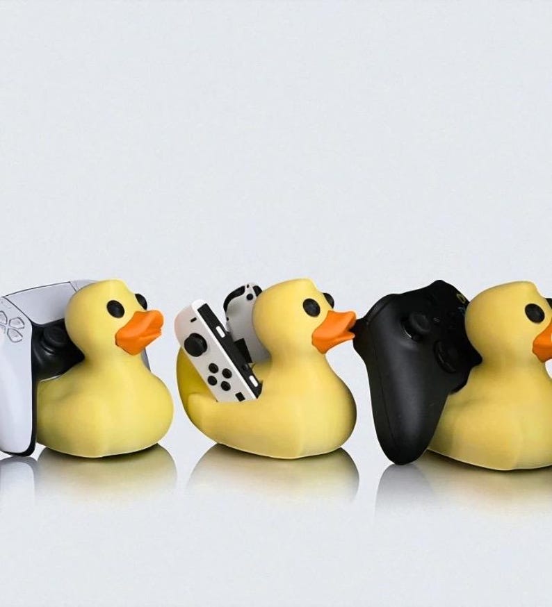 May include: A row of yellow rubber duck figurines, each holding a different video game controller. The controllers are white, black, and a combination of white and black. The ducks have orange beaks and black eyes.