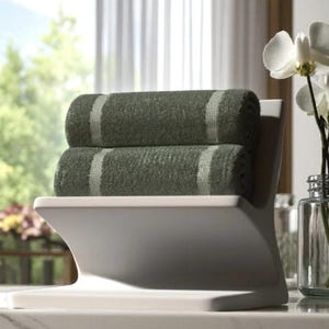 May include: A white towel holder with two rolled green towels. Each towel has a light-coloured stripe down the centre. The holder is on a marble surface, with a window and flowers in the background.