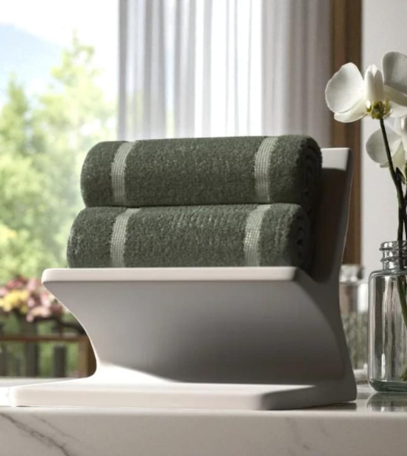 May include: A white towel holder with two rolled green towels. Each towel has a light-coloured stripe down the centre. The holder is on a marble surface, with a window and flowers in the background.