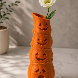 May include: An orange, stacked pumpkin vase with carved jack-o'-lantern faces. A white flower with green leaves is placed in the top opening. The vase is a decorative item for Halloween.
