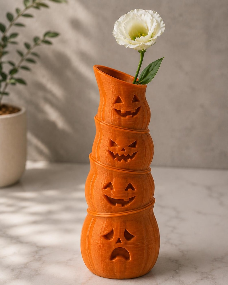 May include: An orange, stacked pumpkin vase with carved jack-o'-lantern faces. A white flower with green leaves is placed in the top opening. The vase is a decorative item for Halloween.