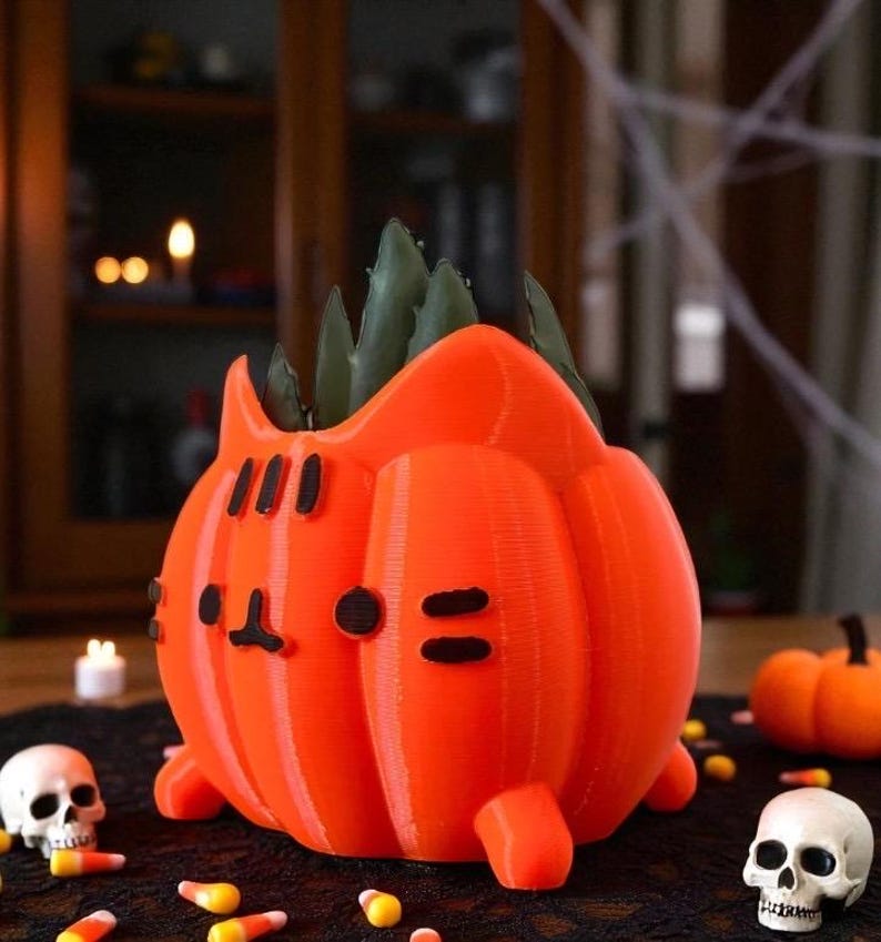 May include: An orange 3D-printed pumpkin-shaped planter with a cat face design. The planter holds a green succulent plant. The image also includes Halloween decorations like candy corn and skull figurines.