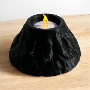 May include: A black, volcano-shaped candle holder with a flickering LED candle. The candle holder has a textured surface and a circular opening to hold the candle. The LED candle has a yellow flame.